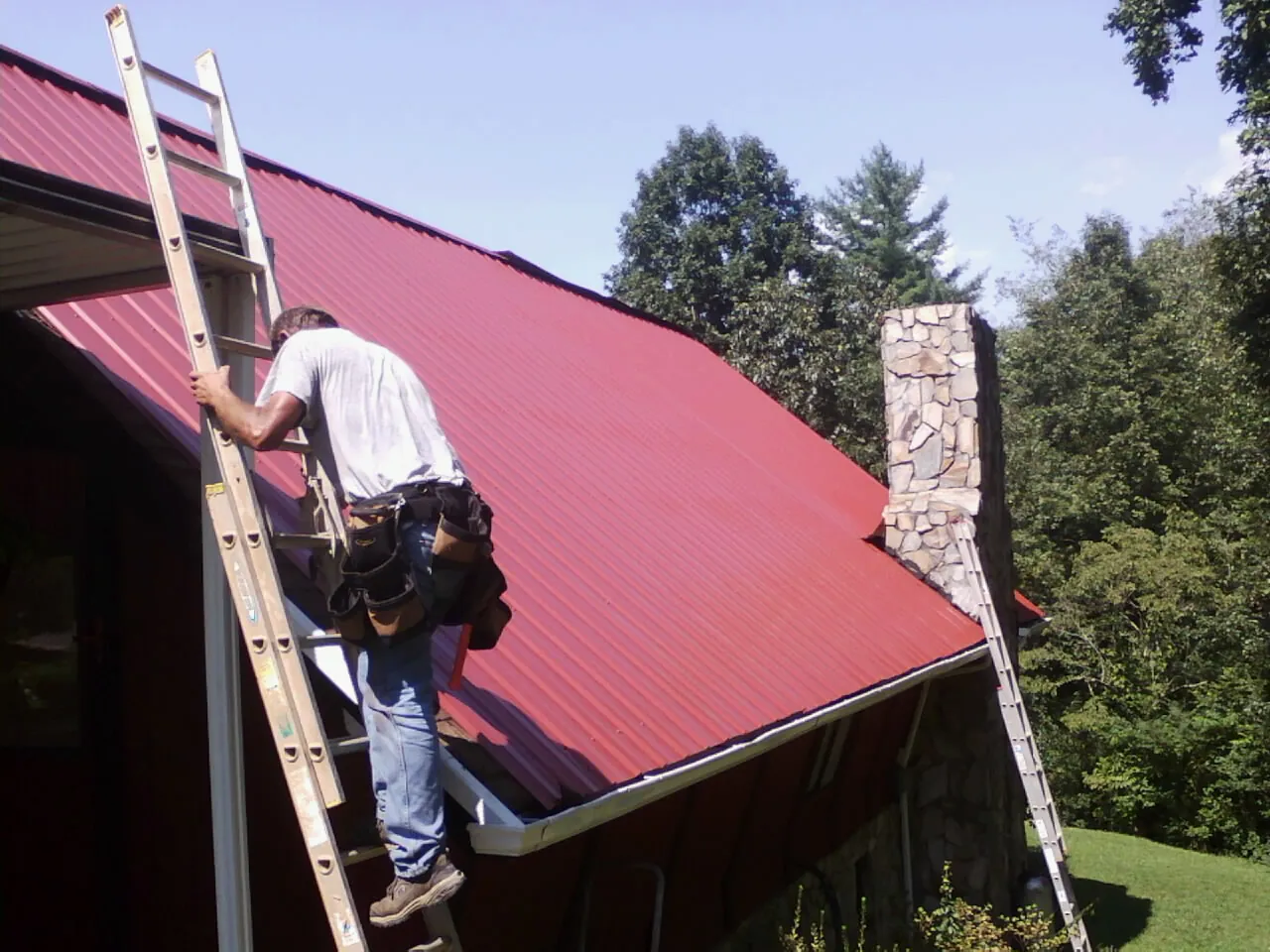 Roofer on a ladder beside a red metal roof and stone chimney — Soffit & Fascia Repair by Roofing Brothers in Pageland